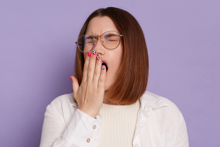 Portrait of sleepy young adult woman wearing white shirt posing isolated over purple background, having sleepless night, yawning, covering mouth with palms, keeps eyes closed.
