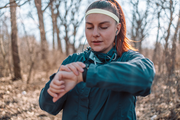 Sport watch run woman checking smartwatch tracker. Trail running runner girl looking at heart rate monitor smart watch in forest wearing jacket sportswear. Female athlete jogger training in woods.