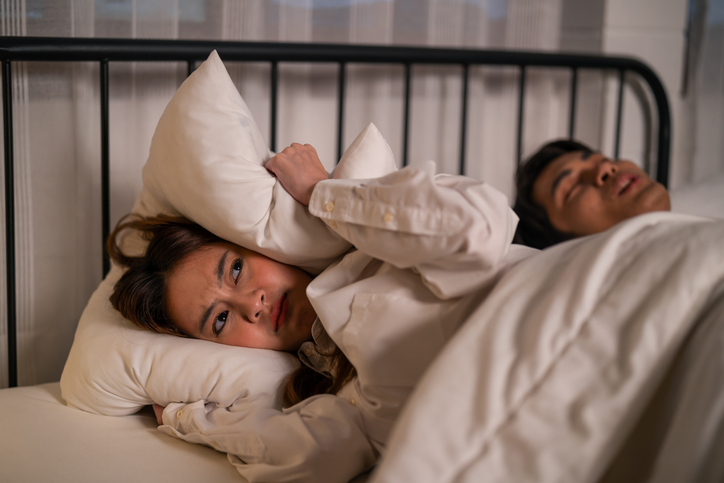 Asian woman feel frustrated from husband snoring while sleep at night. Attractive insomnia angry wife lying down on bed, cannot sleep due to noise and put cozy blanket on her ears in bedroom at home.