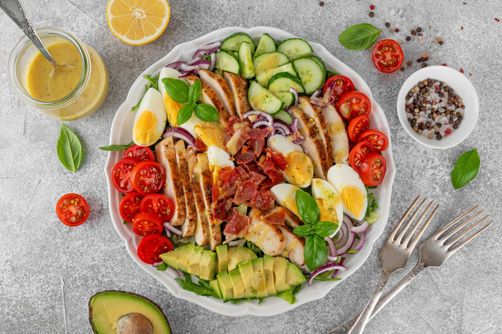 Healthy cobb salad with roasted chicken breast, chopped avocado, tomatoes, bacon, cucumber, hard boiled egg and green leaves on a plate. Top view.