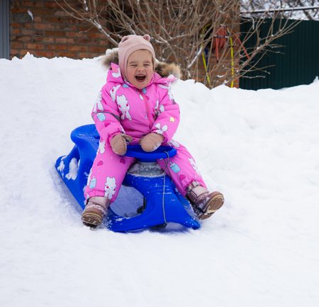 A child sleds down a snowy hill in the backyard, enjoying the winter. Copy Space. In a bright pink jumpsuit, a little girl sleds down a snow hill built by her parents. A fun winter day.