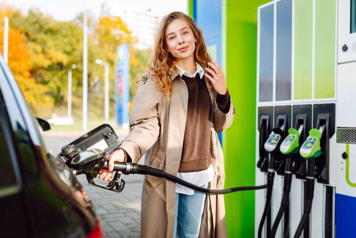 A young woman at a gas station fills up her car with one hand and checks her smartphone with the other.