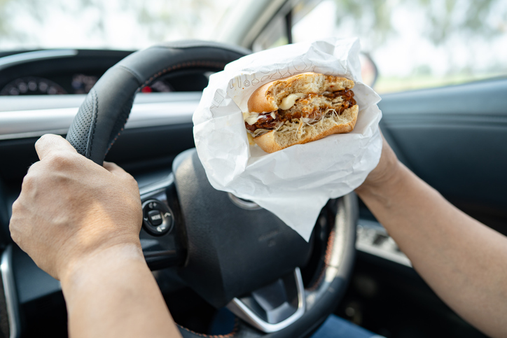 Asian lady holding hamburger to eat in car, dangerous and risk an accident.