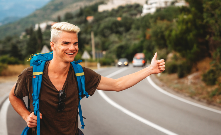 Concept of traveling alone. Young Caucasian man hitchhiking standing by road on sunny summer day