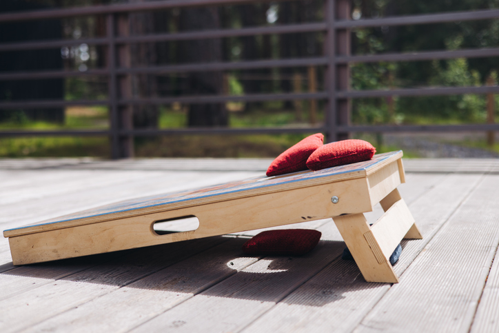 Cornhole game set, process of throwing bean bags, female girl tossing bean sacks, corn hole in the backyard, wooden boards for corn-hole tournament in a summer day on a wooden flooring terrace deck