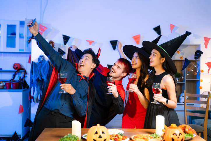 Happy friends in witch and devil costumes celebrating Halloween with drinks, pumpkins, pizza, and laughter at a festive indoor party.