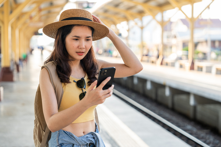 Worried Asian woman checking phone for train delay.